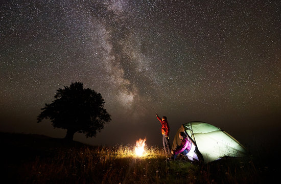 Night camping near silhouette of big tree on grassy valley. Tourist man standing near burning campfire, pointing at beautiful starry sky and Milky way to young woman sitting in glowing tent entrance