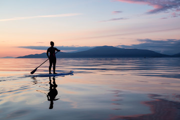 Adventurous girl on a paddle board is paddeling during a bright and vibrant sunset. Taken near...
