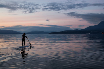 Adventurous girl on a paddle board is paddeling during a bright and vibrant sunset. Taken near Spanish Banks, Vancouver, British Columbia, Canada.