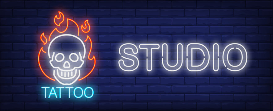 Tattoo Studio Neon Sign. Smiling Scull On Fire And Big Inscription On Dark Brick Wall. Vector Illustration In Neon Style For Tattoo Salon Or Luminous Advertisement