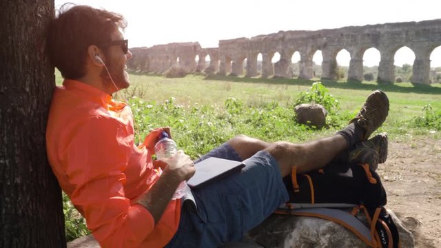 Cyclist with bike sitting on a rock under a tree listening to music with his laptop and drinking water in front of ancient roman aqueduct. Young attractive athletic man with orange sportswear