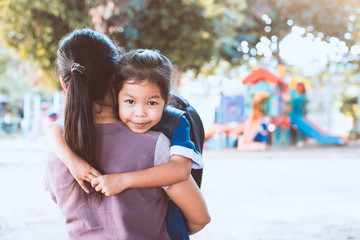 Back to school. Cute asian pupil girl with backpack hugging her mother in the playground before go...