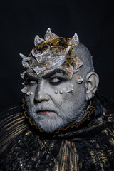 Demon with golden collar on black background. Man with thorns or warts, face covered with glitters. Senior man with white beard dressed like monster. Alien, demon, sorcerer makeup. Fantasy concept.