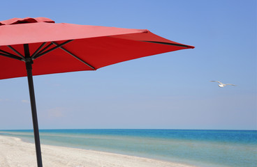 Beach umbrella on a sunny day, sea in background. Beach umbrella close-up. Soaring seagull. Idyllic tropical beach with red umbrella, white sand, turquoise ocean water and blue sky.