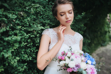 A beautiful and young bride in a lace dress is standing in nature and holding a bouquet in green foliage, in nature. Portrait of a gentle bride with a bouquet.