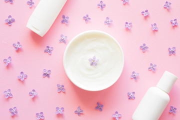 Opened white jar and bottles of natural herbal cream for women on pastel pink background. Purple lilac petals. Care about clean and soft face, hands, legs and body skin. Fresh flowers.