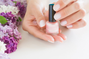 Young, perfect, groomed woman's hands with pink nail varnish bottle. Nails care. Manicure, pedicure beauty salon. Beautiful branches of lilac blossoms on white table. Colorful, fresh flowers.