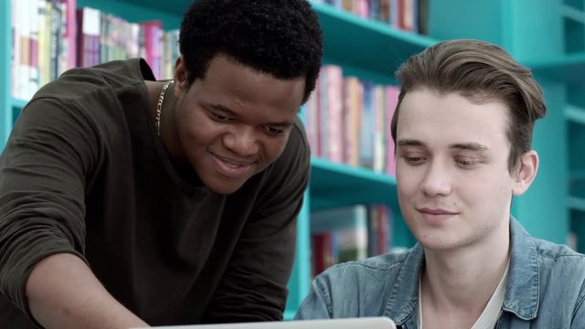 Caucasian college student using laptop to make homework. His cheerful African classmate joining him to help. Boy explaining difficult subject to college friend