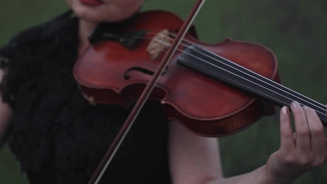 Woman violinist in black lace dress plays the violin on meadow at dusk. Closeup.