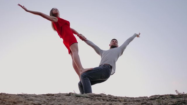 Man and woman practicing yoga together outdoors. Girl in red dress, boy in casual style clothing. Beautiful couple doing acro-yoga on the beach at sunrise, slow motion