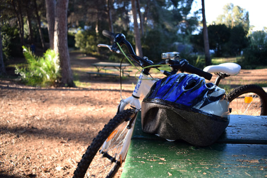 A bicycle standing next to a wooden table in the park on which lies a blue bicycle helmet