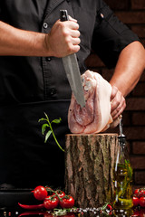 The concept of cooking meat. The man chef, the butcher, cuts raw meat with beef, lamb, veal, holding a knife in his hand, on a wooden table, next to lie raw vegetables, cherry, parsley, dill.