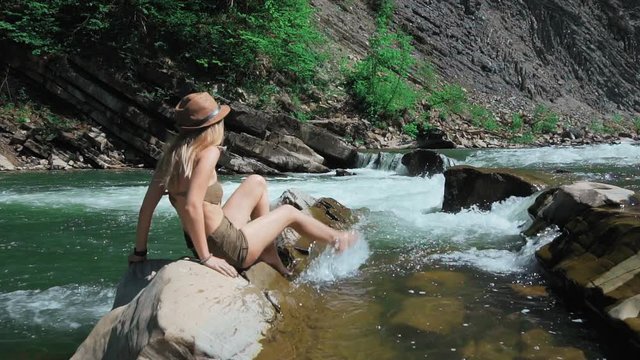 A young woman in a hat, sits on the bank of a mountain river in Switzerland. Moist feet. Fooling around, spray splashing. The concept of freedom and purity. slow motion.