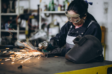Strong and worthy woman doing hard job in car and motorcycle repair shop. She using grinder to fix...