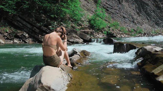 Young woman sitting on rocks on the bank of a mountain river in Switzerland. takes off his hat and spreads his long hair. The concept of freedom and purity, delight. Removal of shackles. Slow motion.