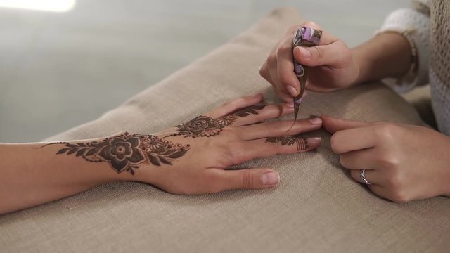 Close-up of hands of artist and customer during mehendi drawing in a salon. Female master is squeezing paste from tube and lying it on skin of girl