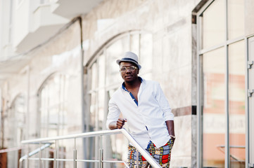 Stylish african american man in white shirt and colored pants with hat and glasses posed outdoor. Black fashionable model boy.