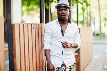 Stylish african american man in white shirt and colored pants with hat posed outdoor. Black fashionable model boy.