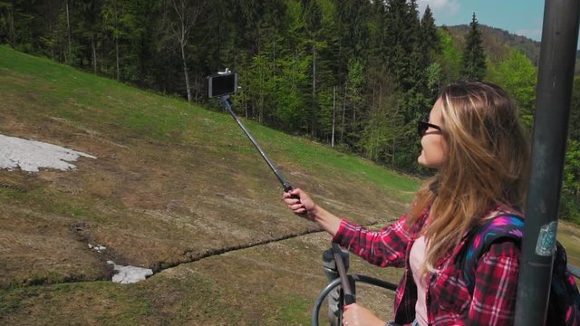 Young woman rides on the mountain lift, Makes selfie using smartphone and Selfiestick. background of the forest in Switzerland. Slow motion.