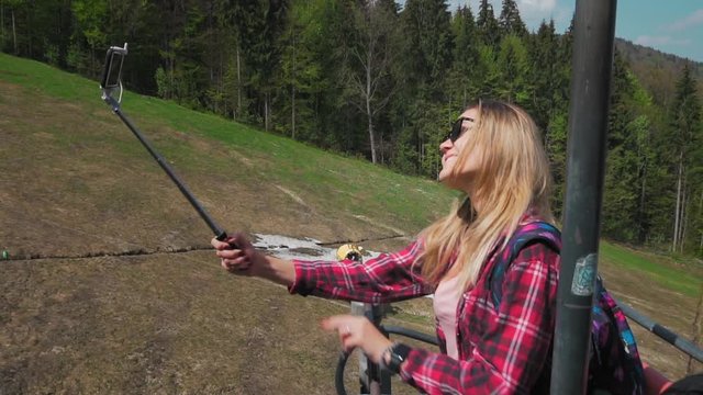 Young woman rides on the mountain lift, Makes selfie using smartphone and Selfiestick. background of the forest in Switzerland. Shows the sign of victory. Slow motion