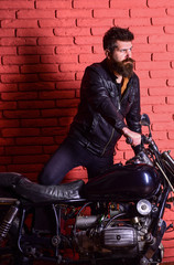 Fototapeta na wymiar Hipster, brutal biker on serious face in leather jacket gets on motorcycle. Masculine passion concept. Man with beard, biker in leather jacket near motor bike in garage, brick wall background.