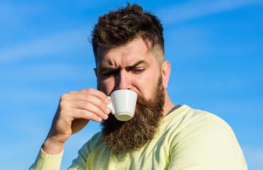 Bearded man with espresso mug, drinks coffee. Coffee gourmet concept. Man with long beard enjoy coffee. Man with beard and mustache on strict face drinks coffee, blue sky background, defocused.