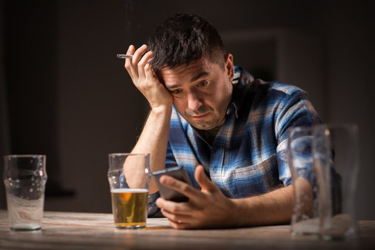 alcoholism, alcohol addiction and people concept - male alcoholic with smartphone drinking beer and smoking cigarette at night