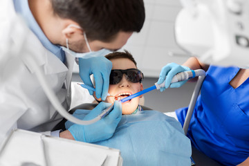 medicine, dentistry and healthcare concept - dentist and assistant with dental drill and saliva ejector treating kid patient teeth at dental clinic