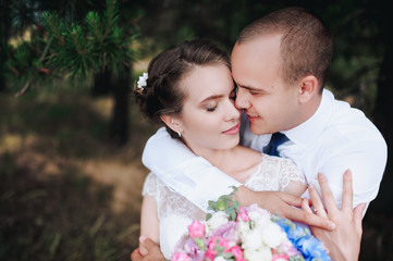 Beautiful and smiling newlyweds gently embrace in a green pine forest on a sunny day. The young groom hugs and kisses from behind a beautiful bride in a lace dress in nature. Wedding portrait close-up