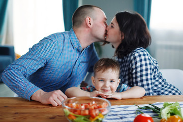 Young husband and wife kiss, son smiling, sitting at the table, family. Concept of a happy family healthy eating