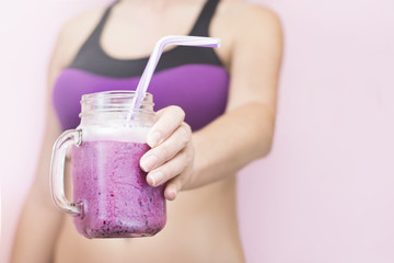 A sporty girl is holding a close-up of a berry smoothie in a glass cup with a straw on a pink background. The concept of a healthy lifestyle and sport.