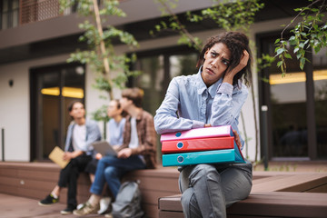 Young upset lady sitting on bench with colorful folders and thoughtfully looking aside while...