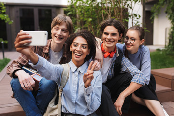Group of young joyful students sitting and taking cute photos on cellphone while happily spending time together in courtyard of university