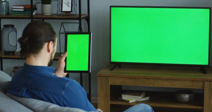 View From Behind On The Caucasian Man Watching TV With Green Screen While Sitting On The Sofa In The Living Room And Holding A Black Tablet Computer With Green Screen Vertically. Focus On TV. Chroma