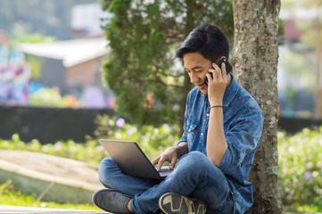 Asian young man with a laptop and smartphone sitting outdoor in a park