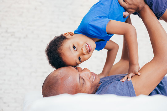 African American family of two, father lifting son up high on bed at home