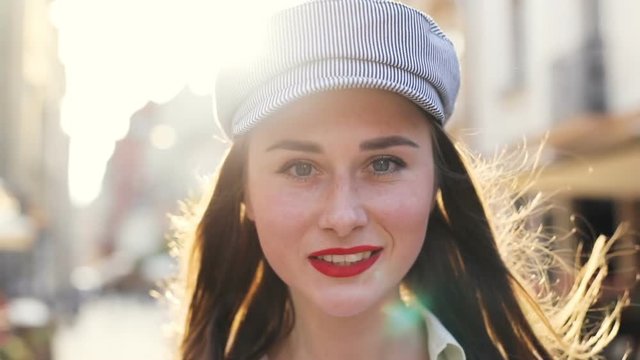 closeup portrait of brunette handsome woman in fashion hat outside smiling and looking at camera with bright sunlight on background city view sunny day light sunray peaceful mood human happiness