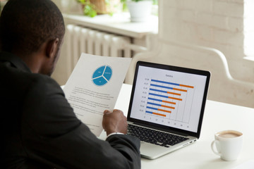 Fototapeta na wymiar African American employee holding handout with presented diagram, comparing financial data with statistics at laptop screen, analyzing business success and raising income. Back view over the shoulder
