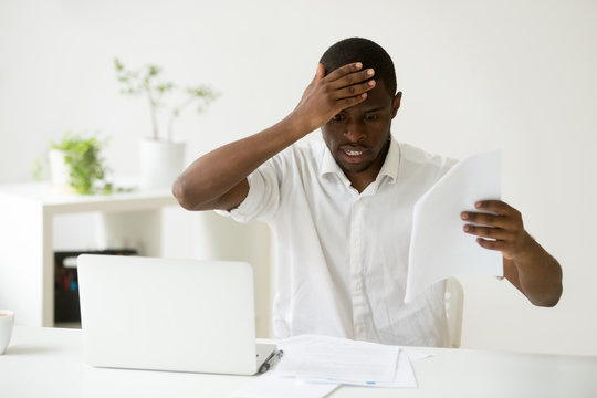 Frustrated African American feeling puzzled looking at printed papers, understanding mistake he made, unsure how to rule the situation out and avoid company business failure, person made redundant