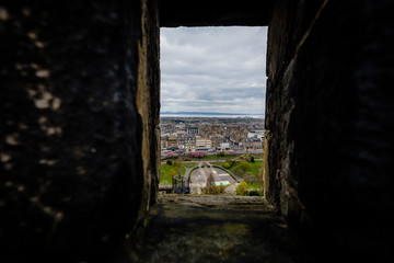 View of Princes Street garden and shops seen through stone windows in Edinburgh Castle during a cloudy day.