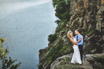 A cute groom hugs a beautiful bride with curly hair, standing on a cliff. Lovely newlyweds stand on a rock, against the background of rocks and the sea.
