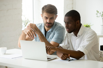 Fototapeta premium Multiethnic work group of two working at laptop pc, discussing developing company strategy, negotiating, brainstorming. Colleague pointing at screen explaining matters to coworker. Diversity concept