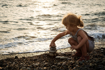 a little girl with long hair is building a pyramid of stones on the beach in the sunset light of the sun