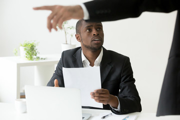 Frustrated shocked African American worker being fired, holding dismissal notice, while Caucasian employer pointing at door asking to leave. Concept of racial discrimination, employment termination