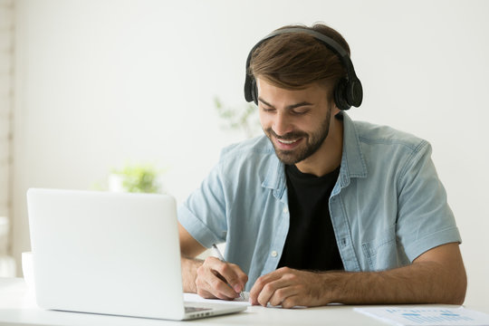Smiling man in headphones watching webinar, listening to web audio course, making notes and writing important information. Happy student enjoying music while taking e-learning class, remote studying