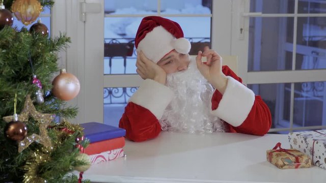 Boring Santa Claus sit near the table and play with pompon of red hat