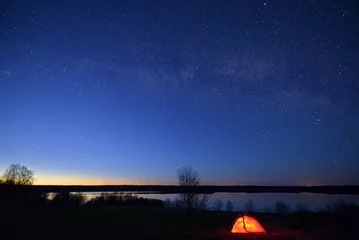 Deurstickers Night landscape with starry sky, Milky Way, lake in the background, and red illuminated tent in the foreground. © SergeG