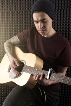A guy with tattoos in a dark T-shirt and hat is playing on the guitar, lit up by the sun.