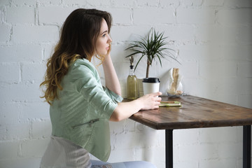 A young girl is sitting at a table with coffee and looking into the distance, propping her head in...