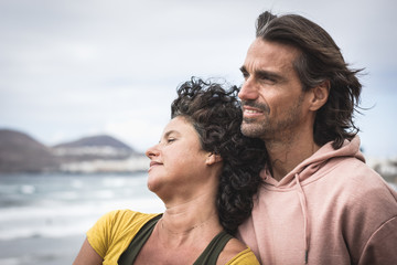 Relaxed couple facing the beach on cloudy, windy day. Young woman leaning on boyfriend's chest while enjoying sea views. Expectant, chill out look concept